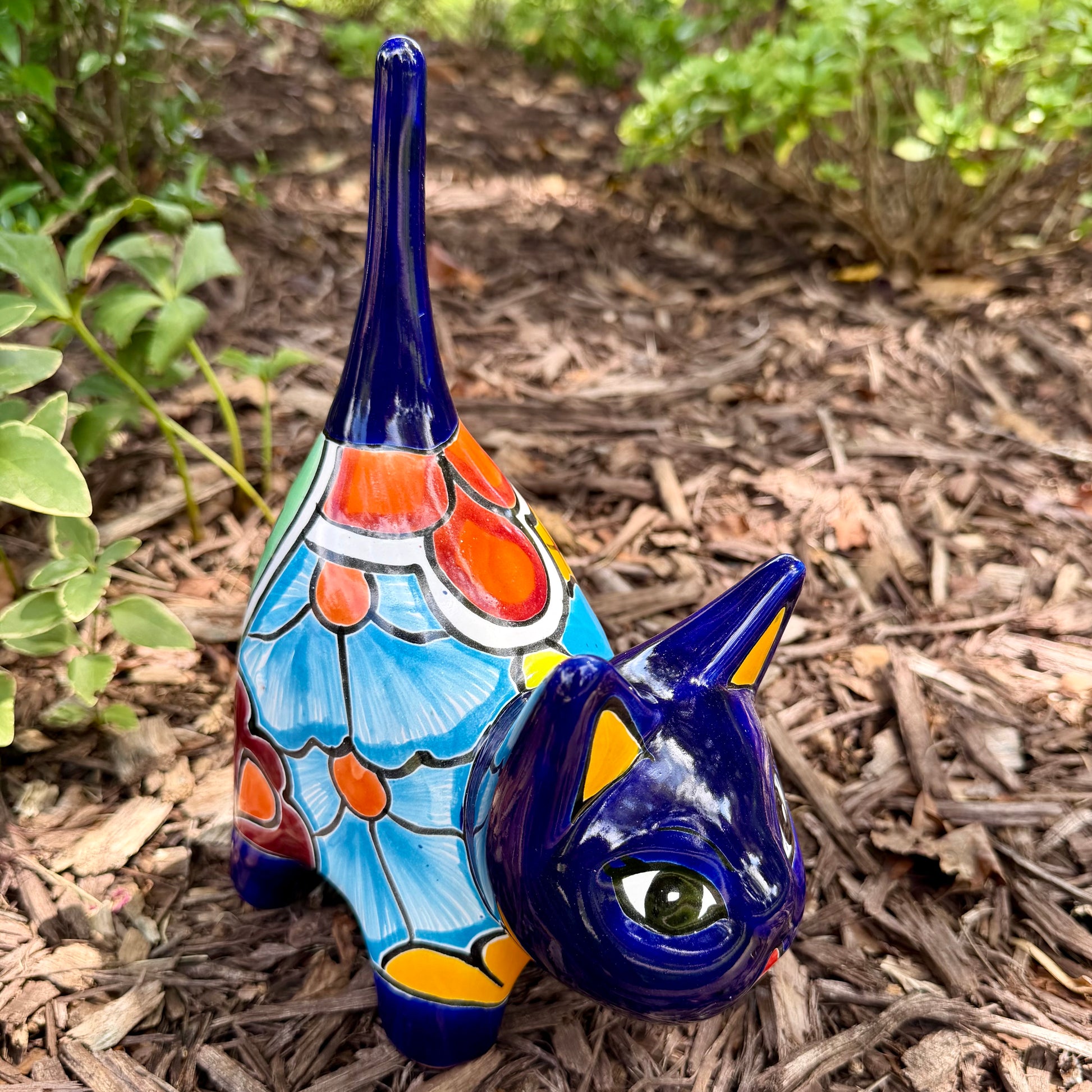 A colorful glazed ceramic cat figurine standing on mulch outdoors, with green plants blurred in the background. The cat is decorated with blue, orange, yellow, and white patterns.