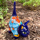 A colorful glazed ceramic cat figurine standing on mulch outdoors, with green plants blurred in the background. The cat is decorated with blue, orange, yellow, and white patterns.
