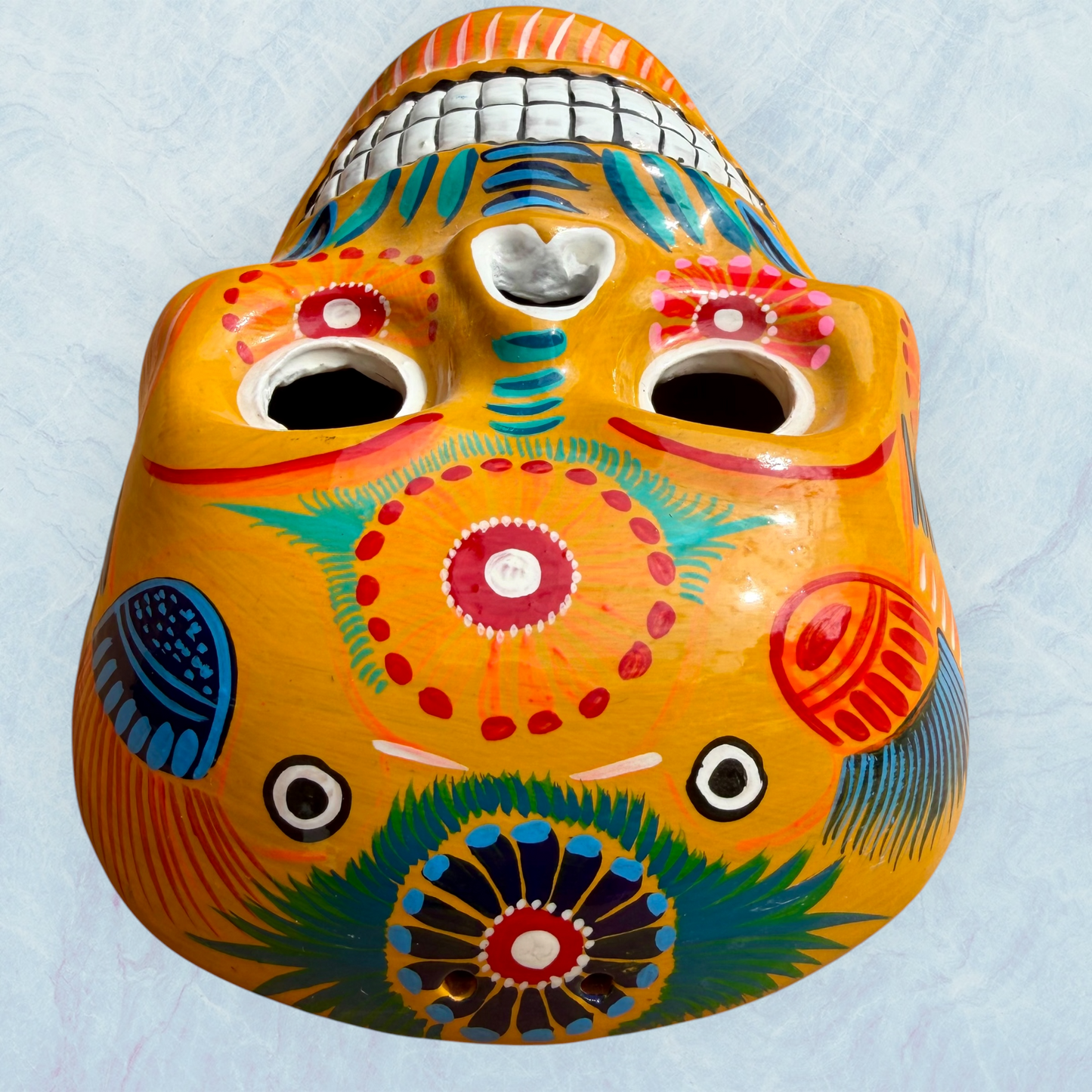 A clay,  golden yellow with floral designs, Mexican-style skeleton skull face mask hanging on a soft blue surface, top forehead view.