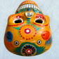 A clay,  golden yellow with floral designs, Mexican-style skeleton skull face mask hanging on a soft blue surface, top forehead view.