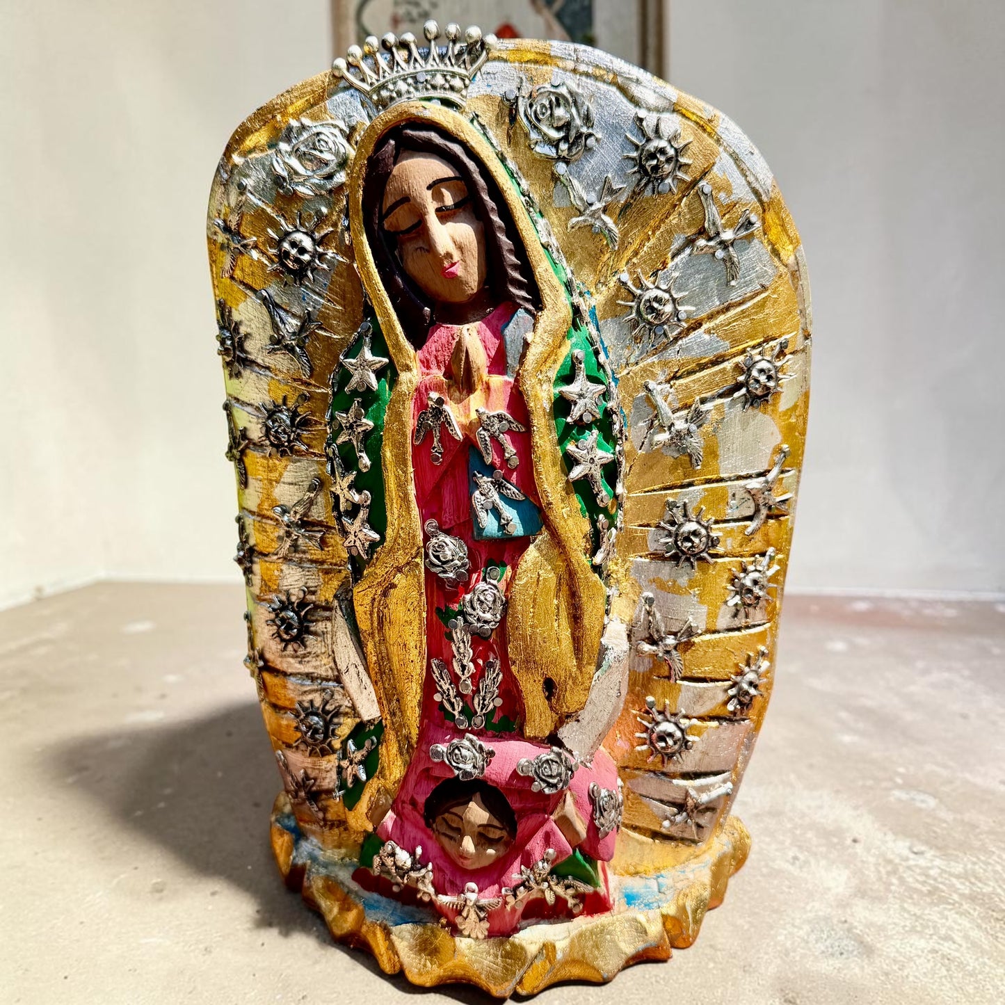 A handmade carved wood Milagros figurine of Our Lady of Guadalupe with a small child at her feet, decorated with metallic silver star and sun milagros charms on a textured gold and brown base.