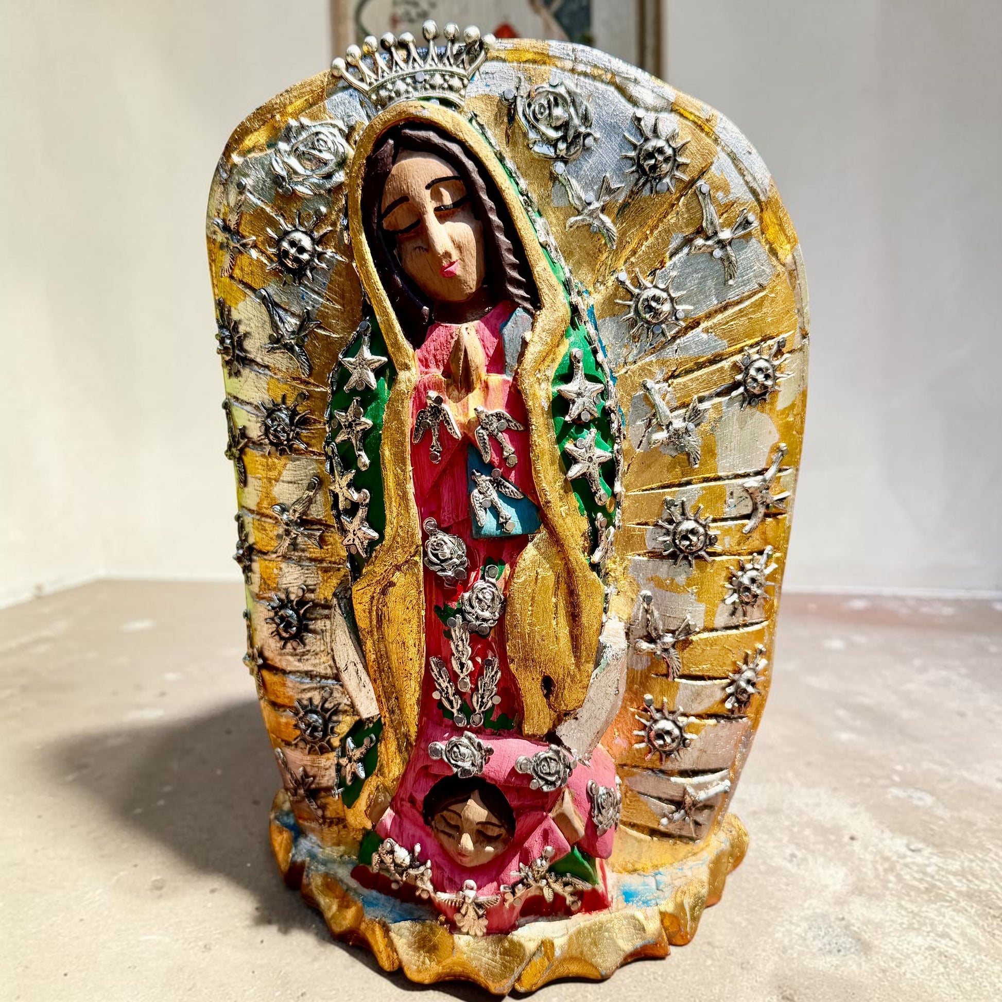 A handmade carved wood Milagros figurine of Our Lady of Guadalupe with a small child at her feet, decorated with metallic silver star and sun milagros charms on a textured gold and brown base.