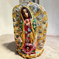 A handmade carved wood Milagros figurine of Our Lady of Guadalupe with a small child at her feet, decorated with metallic silver star and sun milagros charms on a textured gold and brown base.