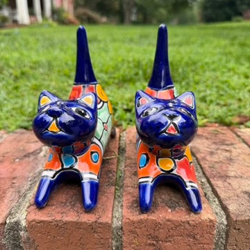 Two cobalt blue with multi-color designs hand-painted talavera ceramic cat figurines resting on a brick ledge with a green grassy lawn front face view.