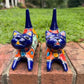 Two cobalt blue with multi-color designs hand-painted talavera ceramic cat figurines resting on a brick ledge with a green grassy lawn front face view.