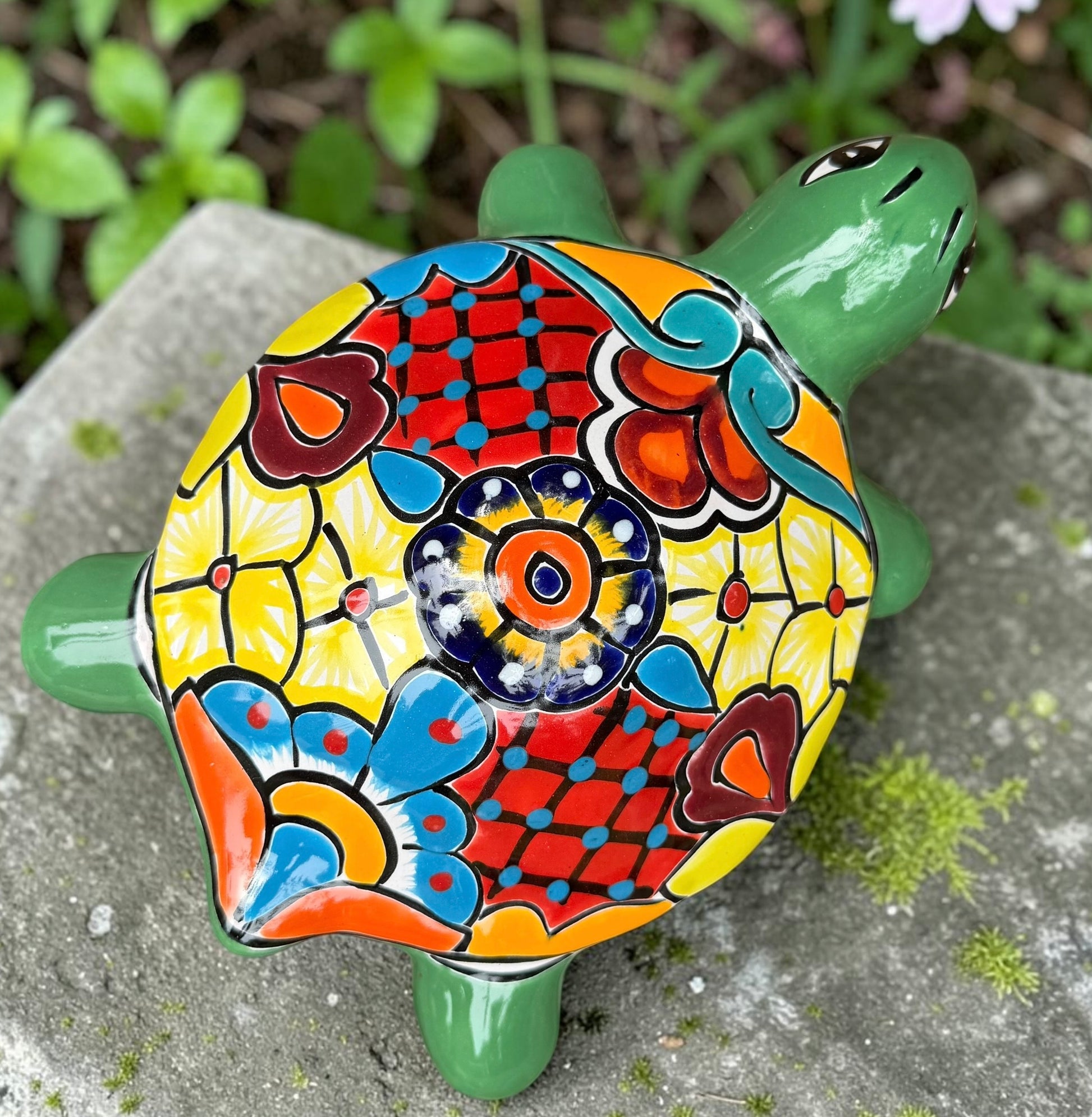 A hand-painted ceramic turtle figurine resting on a gray stone surface with a blurred green garden background. The turtle features a bright green head and legs, and a highly detailed, multicolored shell with yellow, red, orange, blue, and black patterns, top view of Shell.
