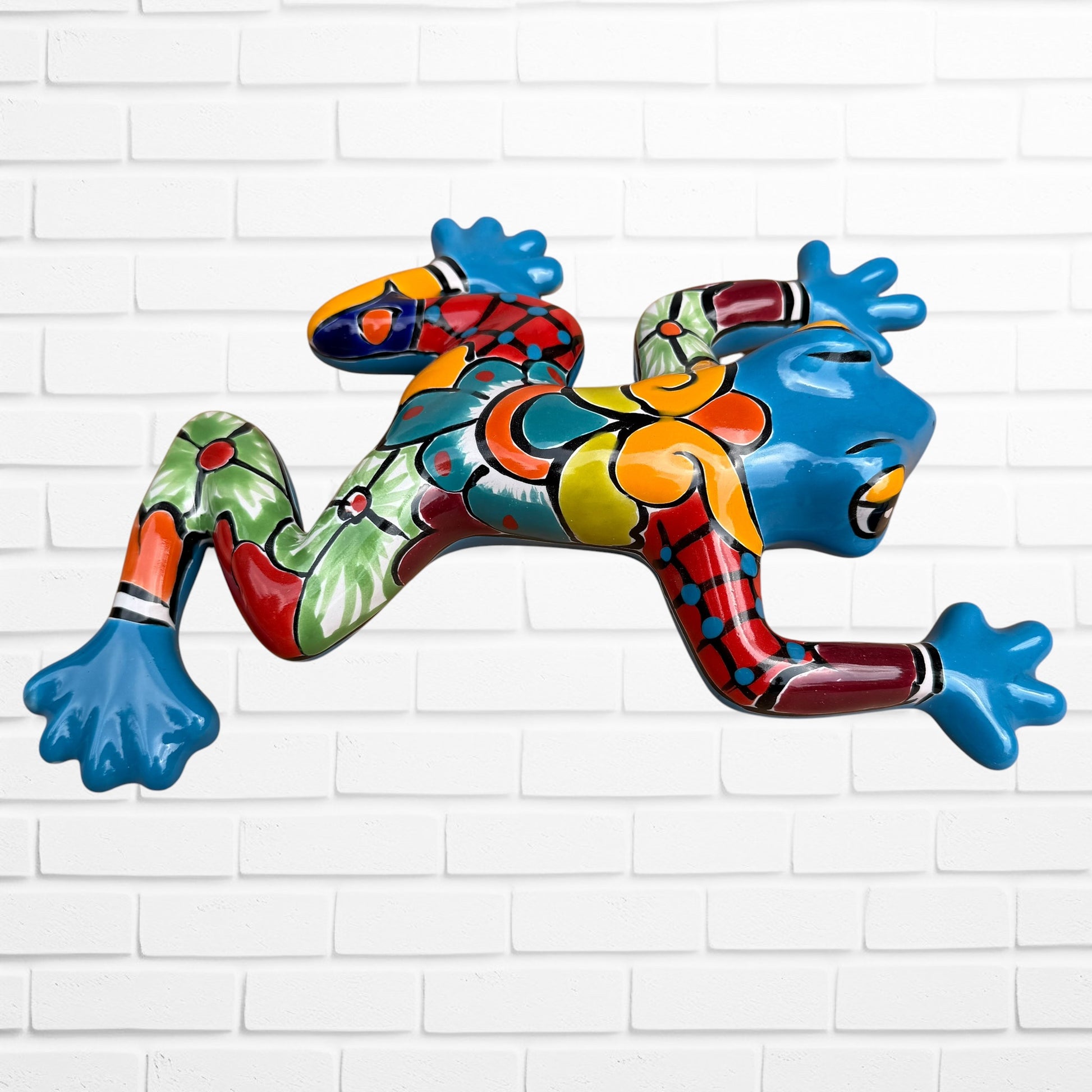 A colorful hand-painted Mexican-style ceramic frog sculpture, shown in a dynamic leaping pose against a white brick wall background.