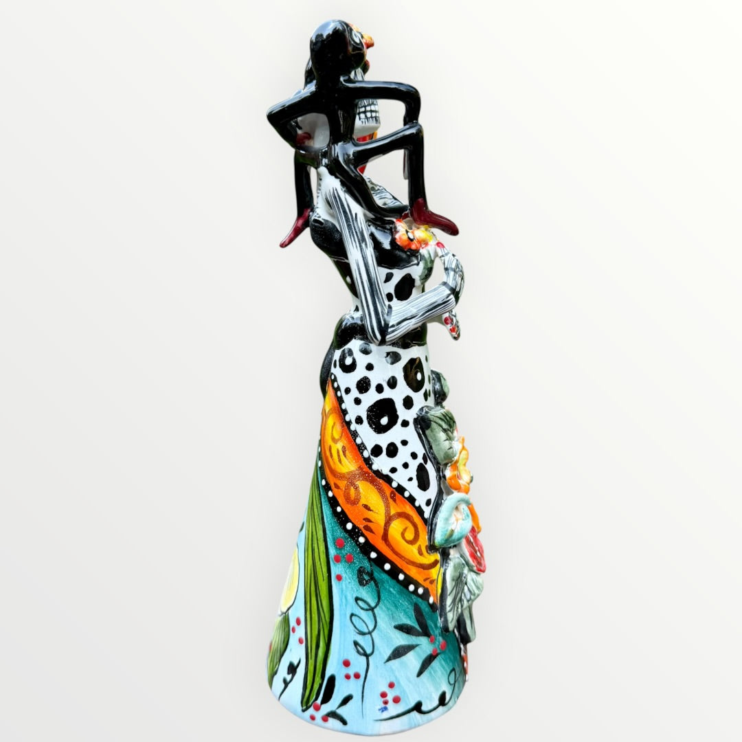 A Day of the Dead Catrina of a Frida skeleton doll holding a black cat, with a red parrot on her shoulder and colorful flowers around the top of her dress. Left side view of the doll.