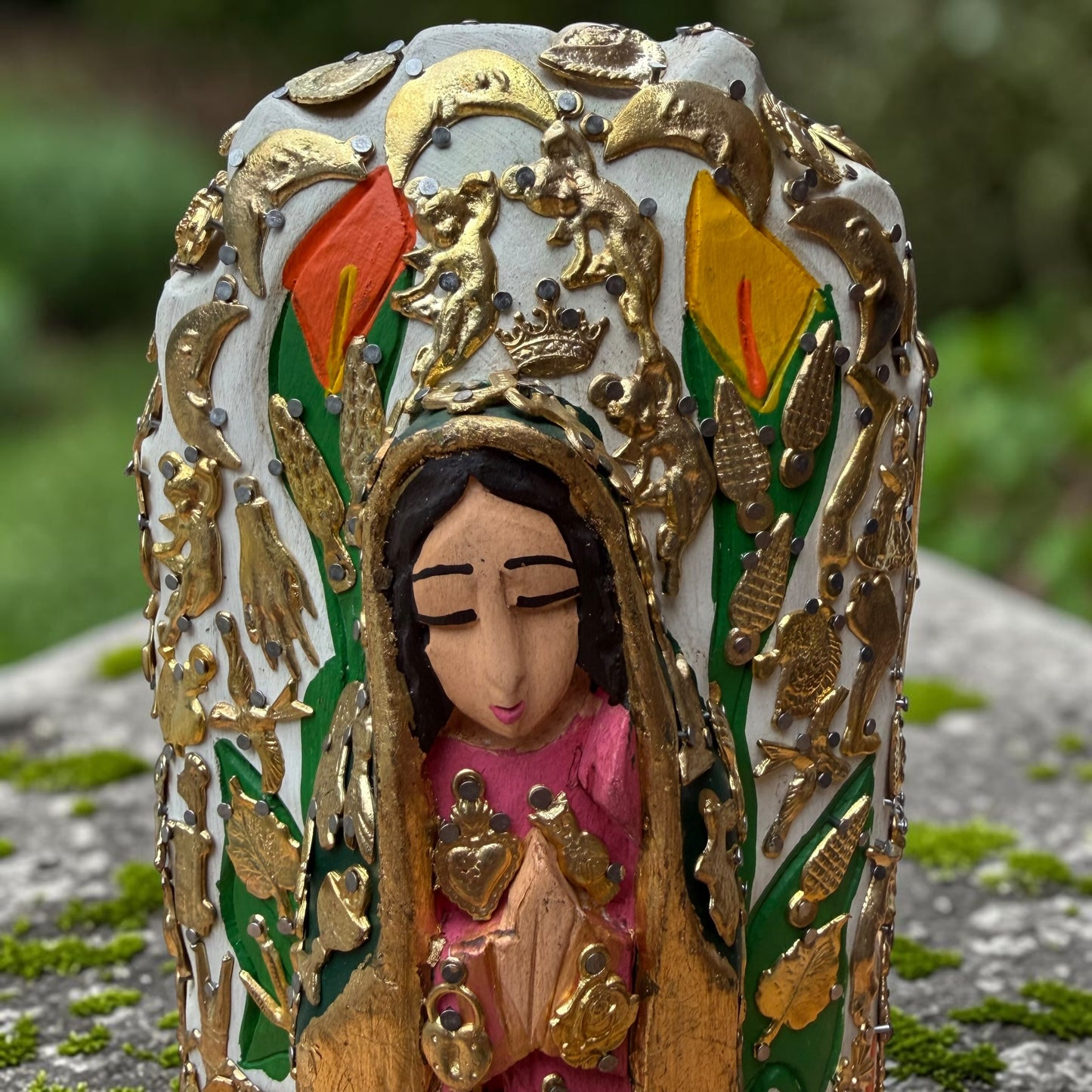 A Milagros religious figurine of Our Lady of Guadalupe with a child, standing on a mossy stone surface with a blurred outdoor garden background. She has a white halo and Lillies adorning the halo. Face close up.