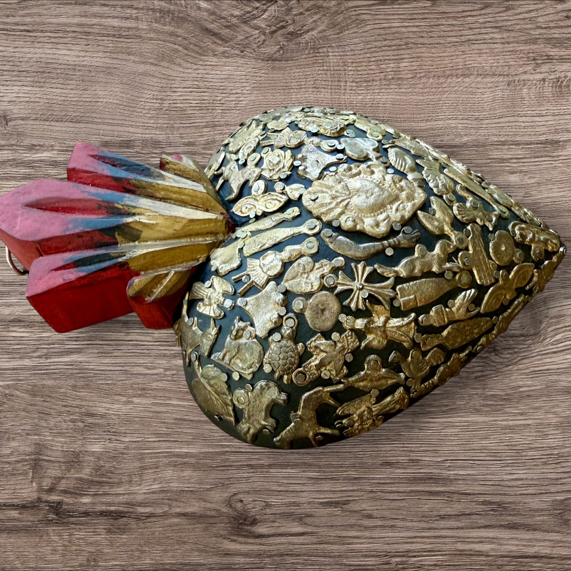 A Mexican Milagros large heart in black with a gold and red flaming top, covered in antiqued charms, left side view.
