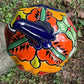 A hand-painted ceramic jack-o'-lantern pumpkin ornament with a bright orange face, red carved eyes and mouth, a blue stem, and colorful side patterns, sitting outdoors on a stone pedestal in a grassy yard top view.