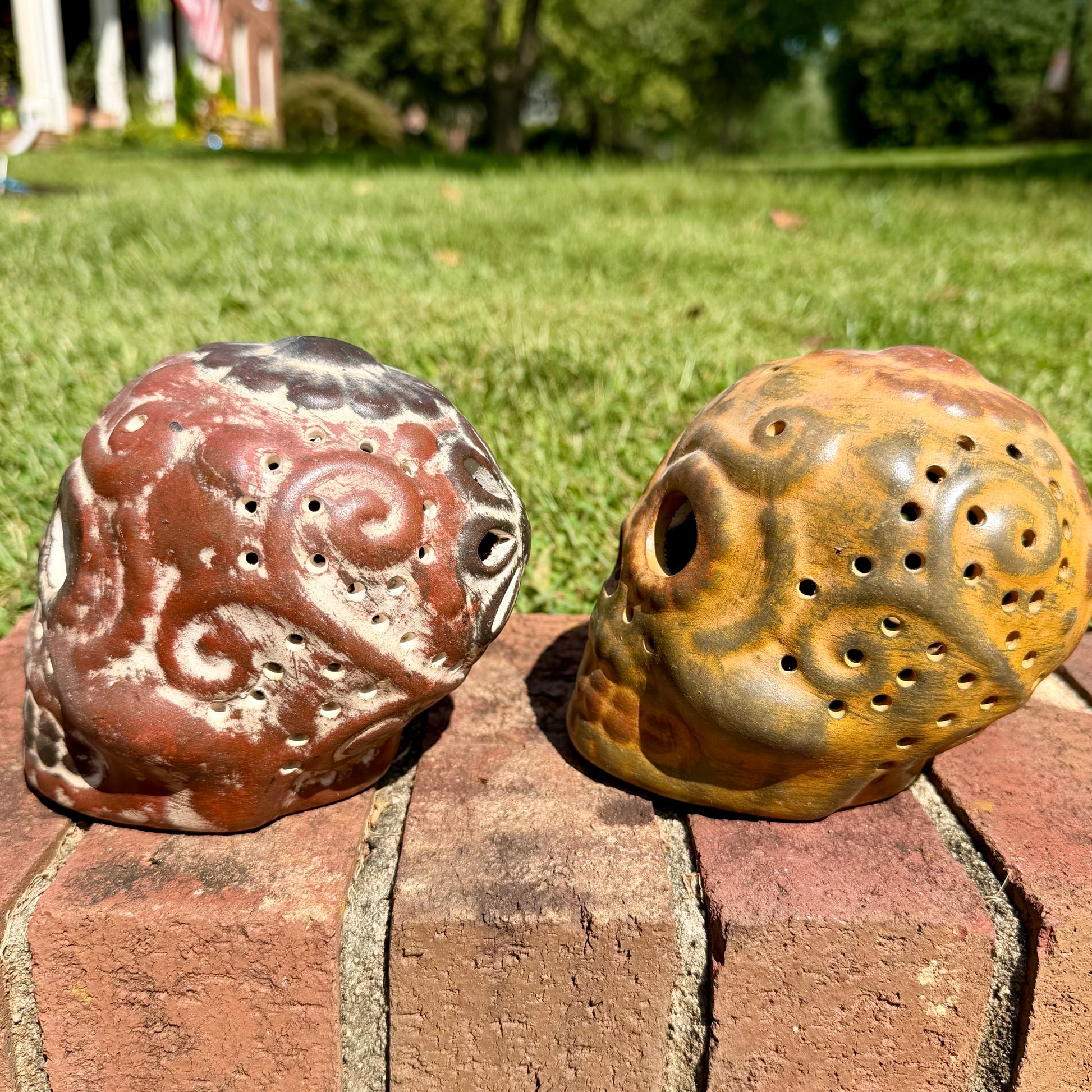 Two decorative clay sugar skull ornaments with Day of the Dead style painted patterns, displayed side-by-side on a brick surface outdoors with a grassy lawn and trees in the background, left side view.