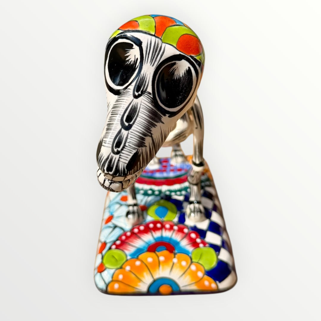 A talavera catrina dog skeleton figurine with colorful hand-painted floral and geometric patterns, standing on a matching decorated base against a plain white background. Front face view.