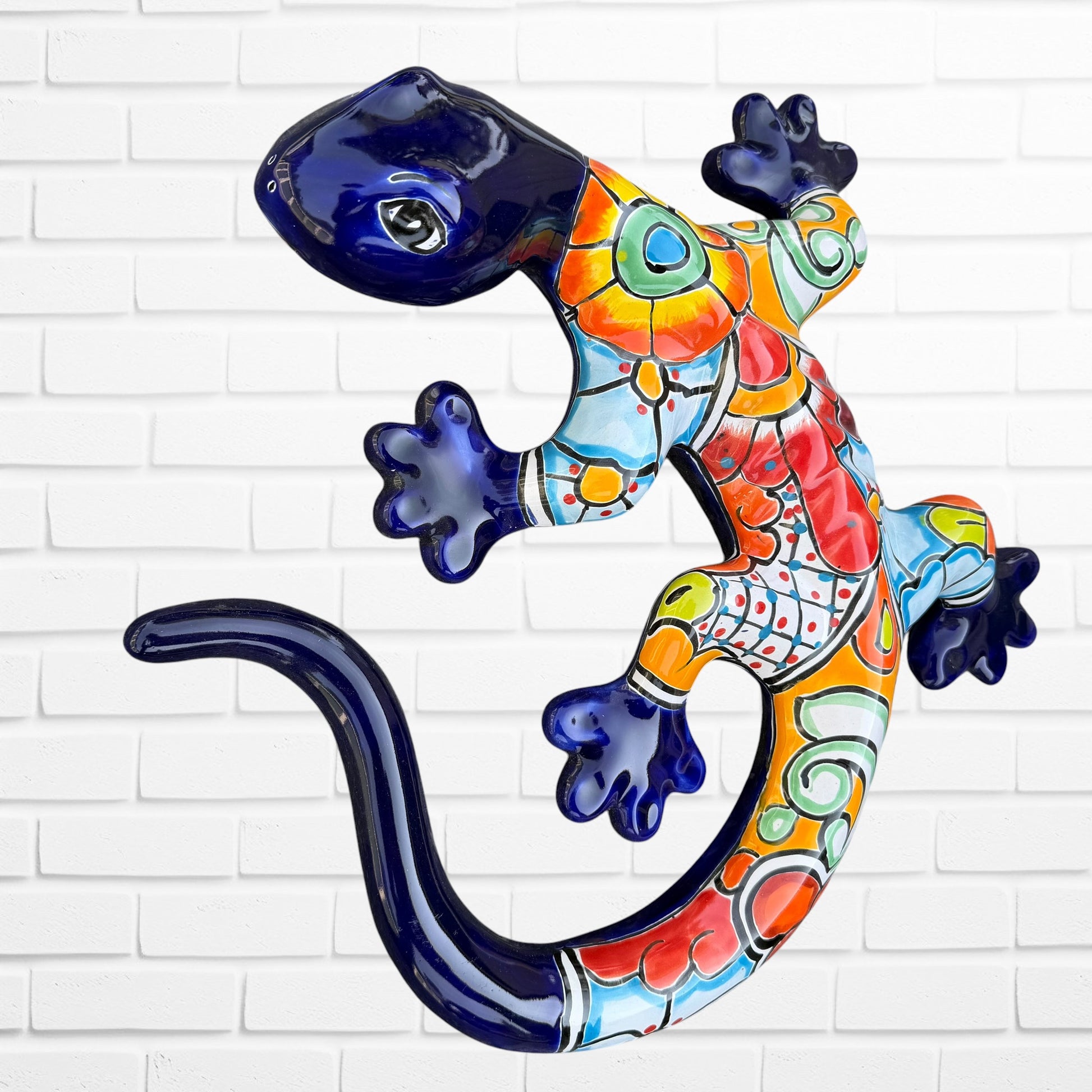 A colorful Talavera ceramic wall sculpture of a salamander, featuring a glossy finish and intricate hand-painted patterns in blue, orange, red, yellow, and white, mounted against a white brick wall.