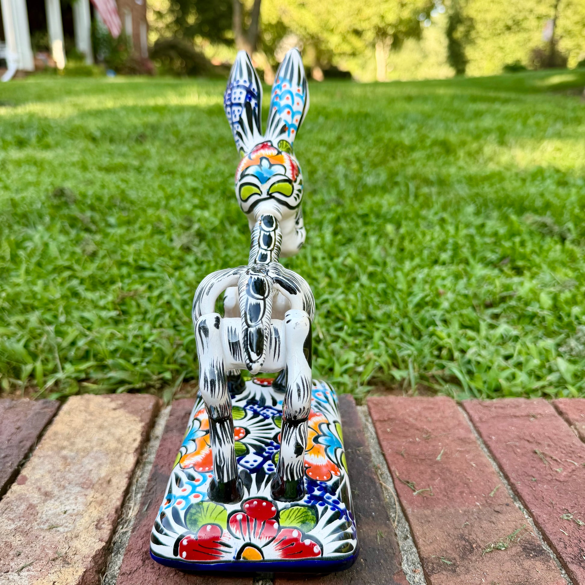 A colorful ceramic donkey Catrina figurine with a skeletal white-and-black design and a floral base, photographed outdoors on a brick surface with a blurred green lawn and house in the background. This is the back view with the tail.
