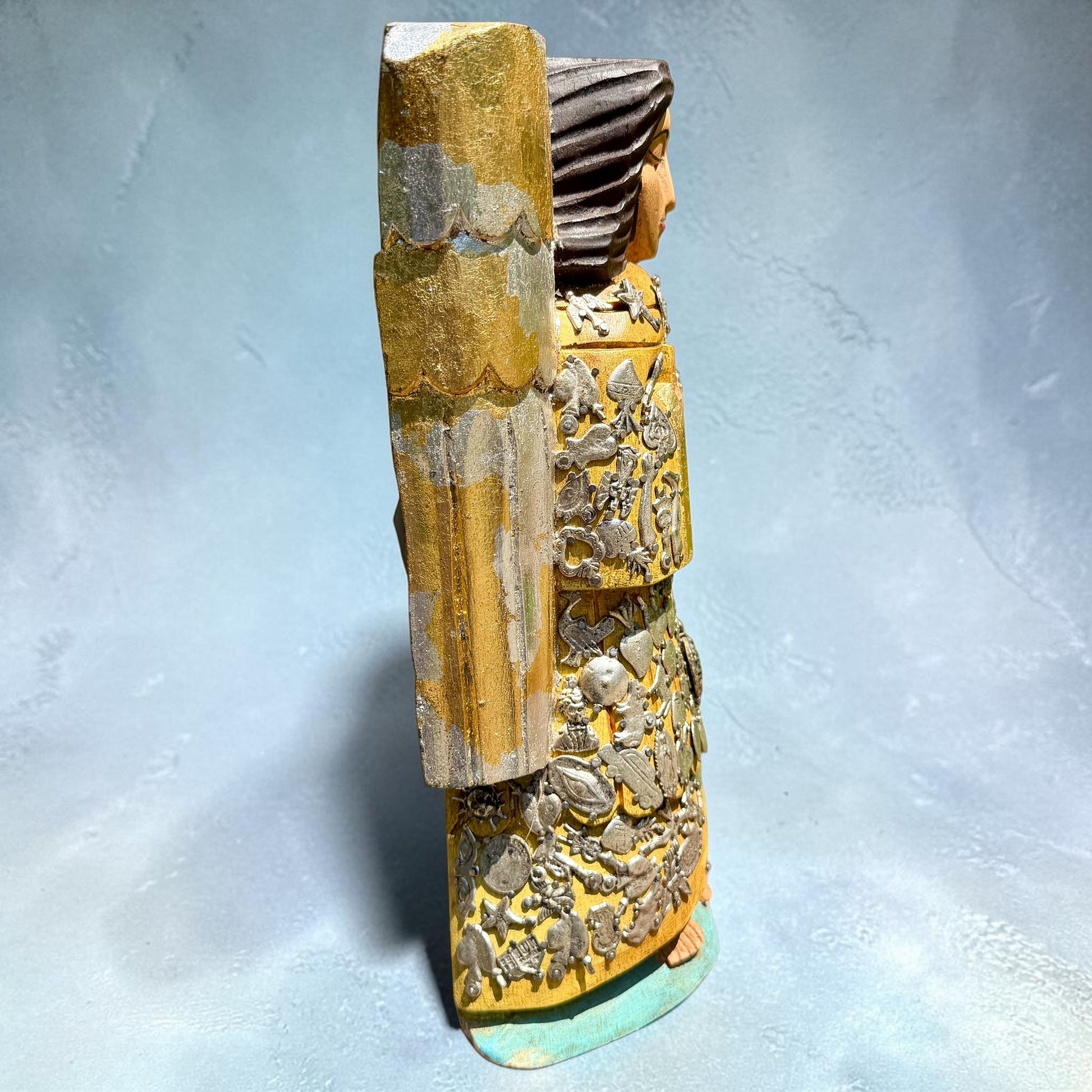 Milagros wooden guardian angel figurine wearing a yellow robe with gold and silver metallic details, standing on a light blue textured background, left side view.