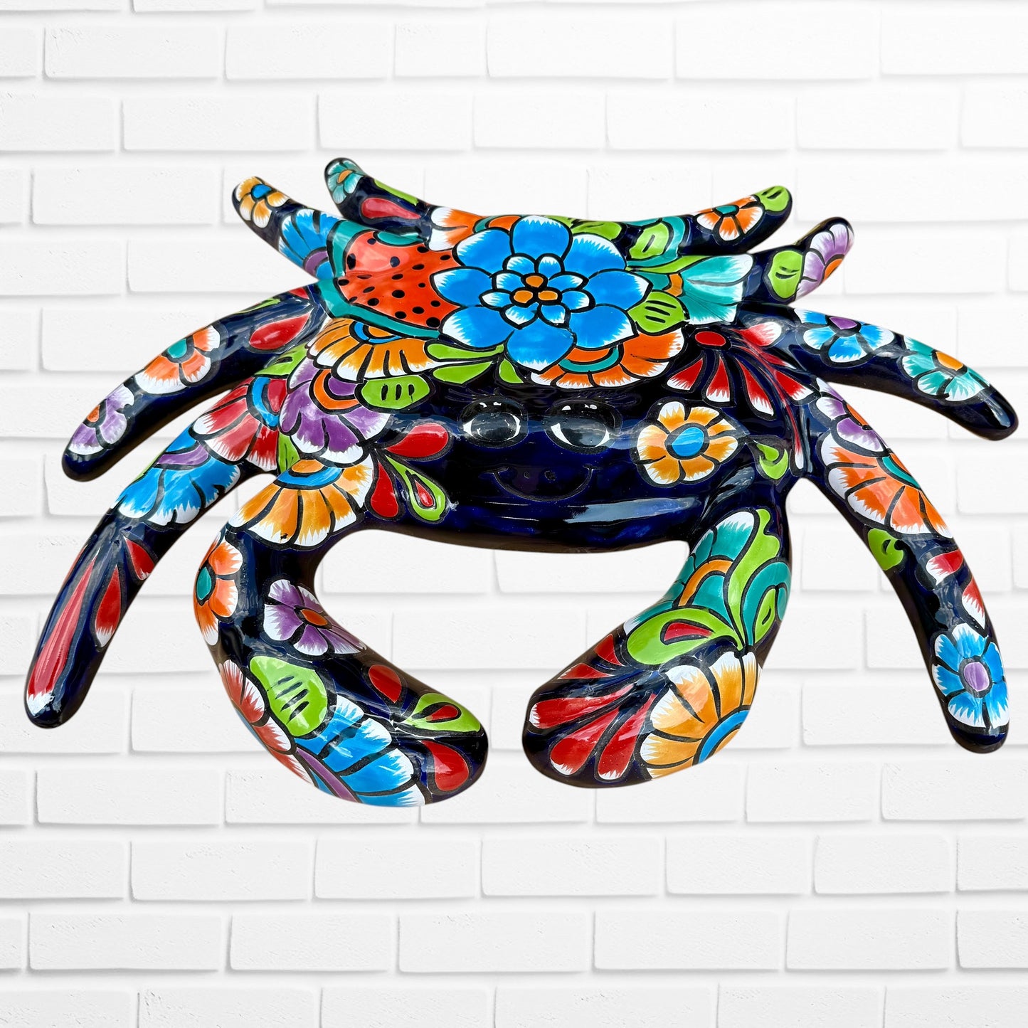 A colorful talavera crab wall art sculpture decorated with vibrant floral patterns, shown against a white brick wall background, Face view of Crab.