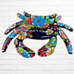 A colorful talavera crab wall art sculpture decorated with vibrant floral patterns, shown against a white brick wall background, Face view of Crab.