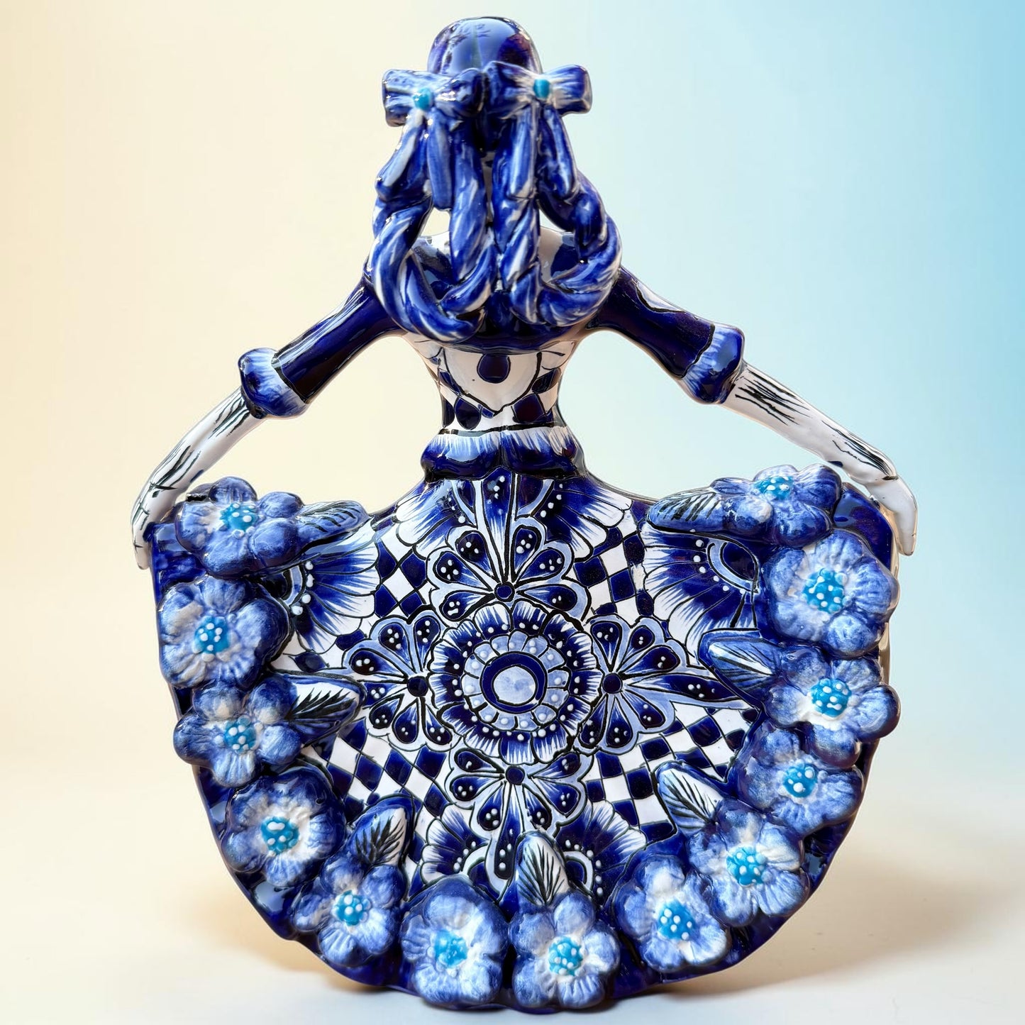 A Day of the Dead style talavera figurine of a skeleton woman in a wide floral skirt, painted in deep blue, black, white, and light blue on a soft beige-to-blue gradient background. Back view of ballerina.