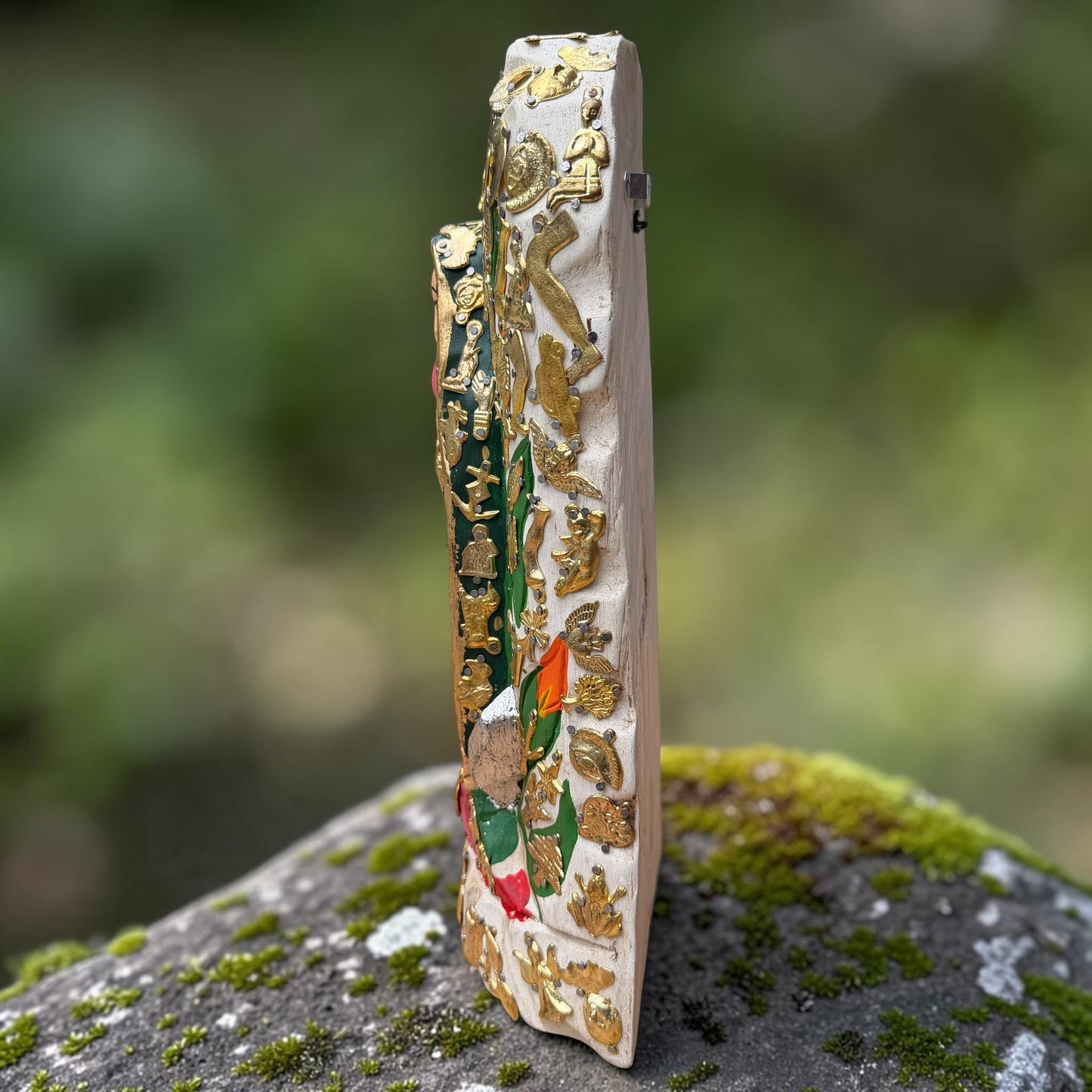 A Milagros religious figurine of Our Lady of Guadalupe with a child, standing on a mossy stone surface with a blurred outdoor garden background. She has a white halo and Lillies adorning the halo. Right side view of Milagros.