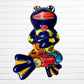 A colorful ceramic frog figurine sitting with arms crossed, decorated with vibrant hand-painted patterns in blue, yellow, red, orange, and light blue, set against a white brick wall background.