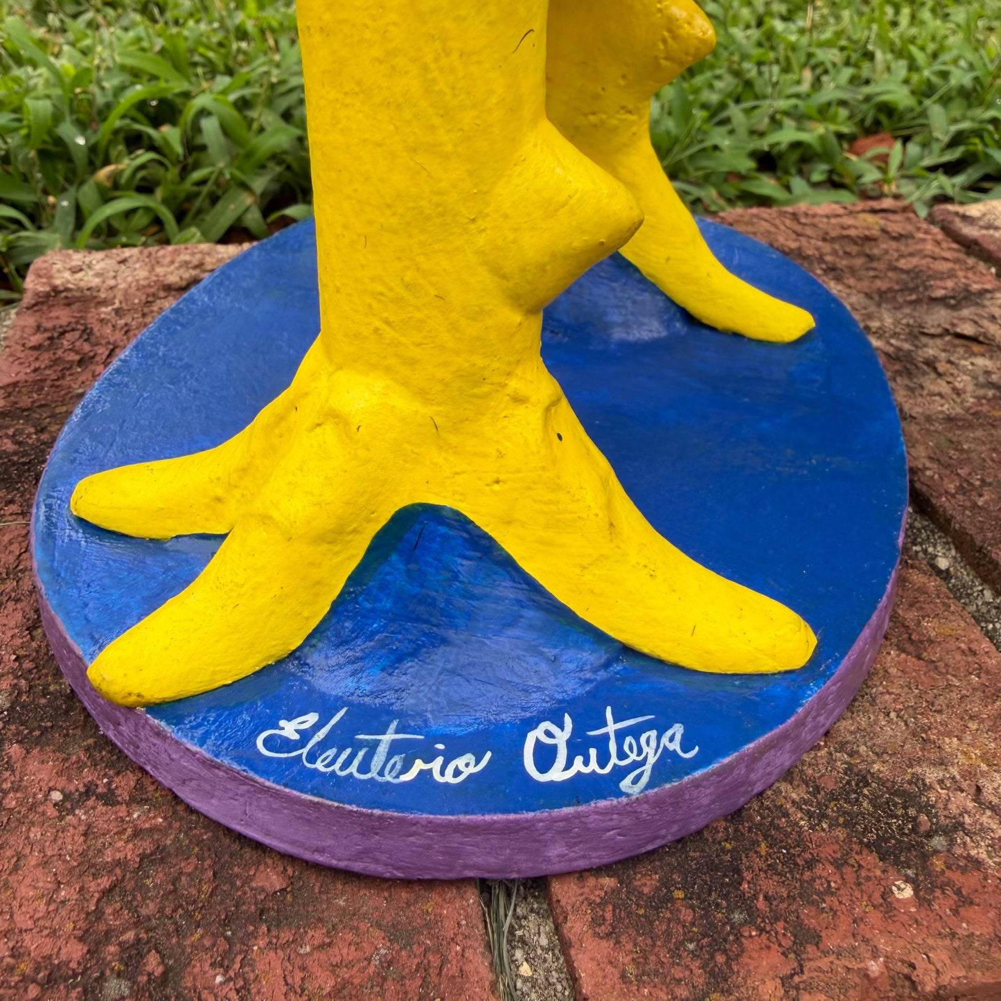 A colorful decorative rooster figurine standing on a brick surface outdoors, with a grassy lawn and trees in the background. The rooster is primarily red and yellow with many small multicolored flowers and painted details across its body and tail. This photo shows the artist signature.