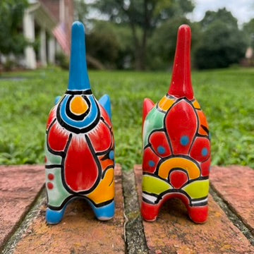 Two hand-painted Mexican-style ceramic cat figurines, one blue and one red, decorated with colorful geometric patterns, standing on a brick surface outdoors back tail view