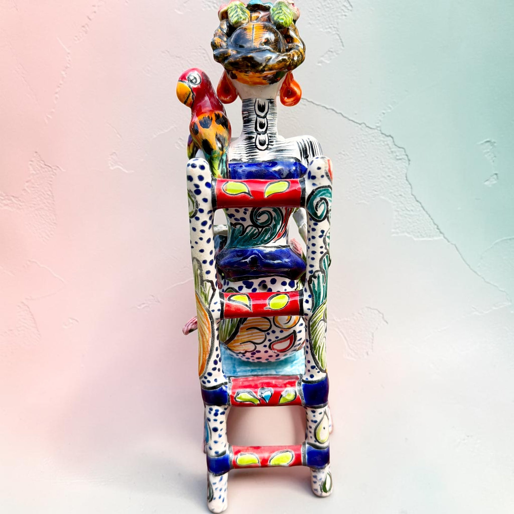 Frida Kahlo Talavera day of the dead figurine sitting in a chair holding a monkey with a parrot on her shoulder, back side view.