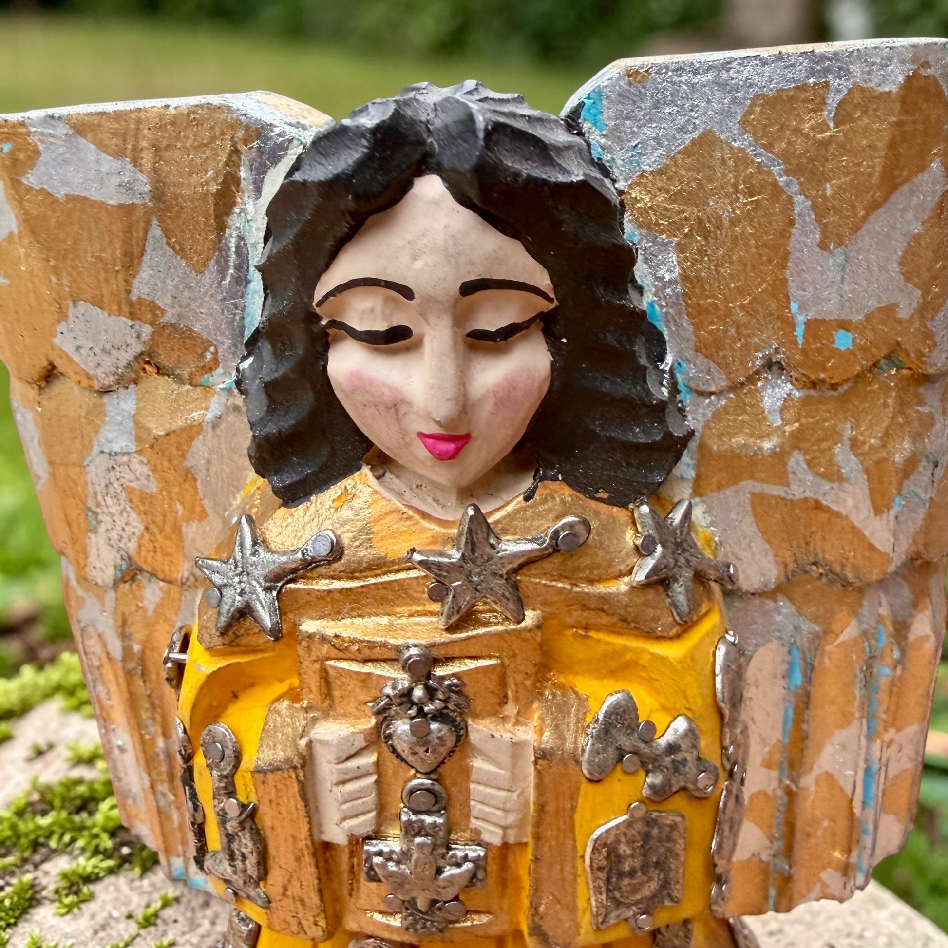 A handcrafted wooden Milagros angel figurine with large wings, decorated with metallic silver charms and painted in yellow, and orange, standing on a mossy stone with a blurred green garden background. Face close up.