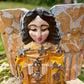 A handcrafted wooden Milagros angel figurine with large wings, decorated with metallic silver charms and painted in yellow, and orange, standing on a mossy stone with a blurred green garden background. Face close up.