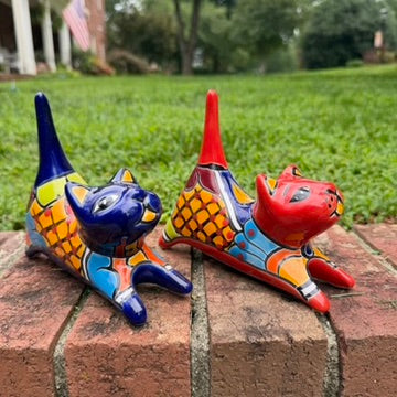 Two colorful ceramic cat figurines, one blue and one red, decorated with geometric patterns, displayed on a brick ledge with a green lawn in the background.