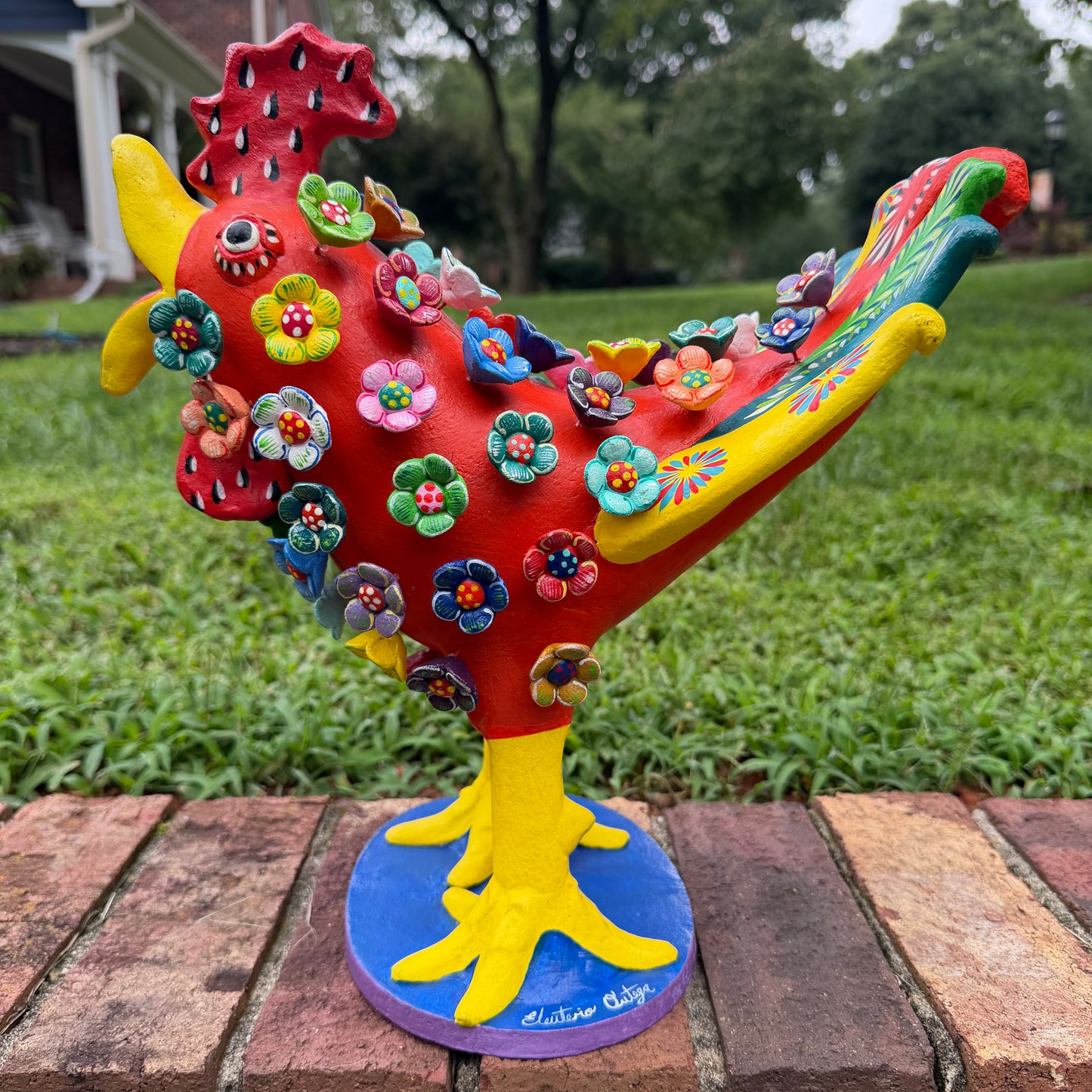 A colorful decorative rooster figurine standing on a brick surface outdoors, right side view, with a grassy lawn and trees in the background. The rooster is primarily red and yellow with many small multicolored flowers and painted details across its body and tail.