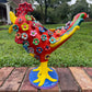 A colorful decorative rooster figurine standing on a brick surface outdoors, right side view, with a grassy lawn and trees in the background. The rooster is primarily red and yellow with many small multicolored flowers and painted details across its body and tail.