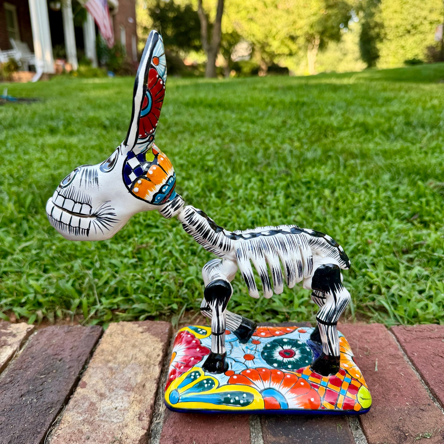 A colorful hand-painted Talavera Catrina donkey figurine, standing on a colorful floral base in an outdoor backyard setting, right side view of donkey.