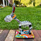 A colorful hand-painted Talavera Catrina donkey figurine, standing on a colorful floral base in an outdoor backyard setting, right side view of donkey.