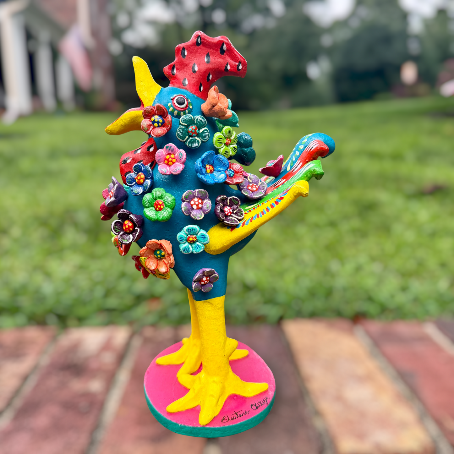A colorful decorative rooster folk art sculpture with a blue body, yellow legs and beak, and many bright flower ornaments, standing on a pink circular base outdoors on a brick path with a blurred green lawn behind it. Rooster right side view. 