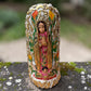 A Milagros religious figurine of Our Lady of Guadalupe with a child, standing on a mossy stone surface with a blurred outdoor garden background. She has a white halo and Lillies adorning the halo.