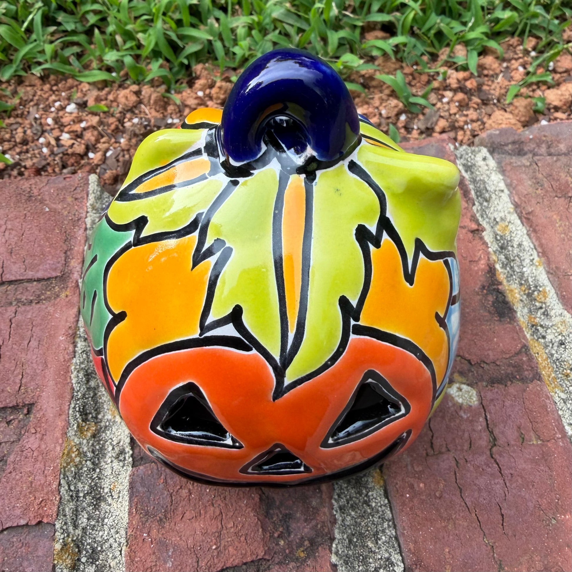 One hand-painted Talavera ceramic jack-o'-lantern pumpkins with orange faces and yellow, blue, and red green designs, sitting on a brick path in a grassy backyard, top stem view.