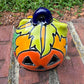 One hand-painted Talavera ceramic jack-o'-lantern pumpkins with orange faces and yellow, blue, and red green designs, sitting on a brick path in a grassy backyard, top stem view.