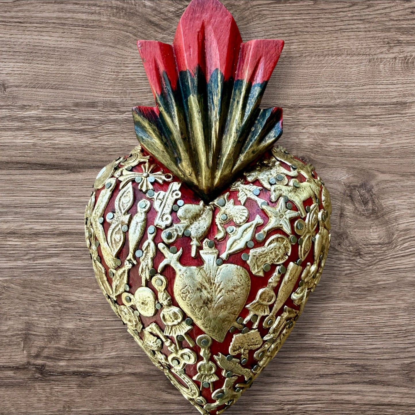 A Mexican Milagros medium heart in red with a gold and red flaming top, covered in antiqued golden charms. 
