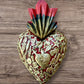 A Mexican Milagros medium heart in red with a gold and red flaming top, covered in antiqued golden charms. 