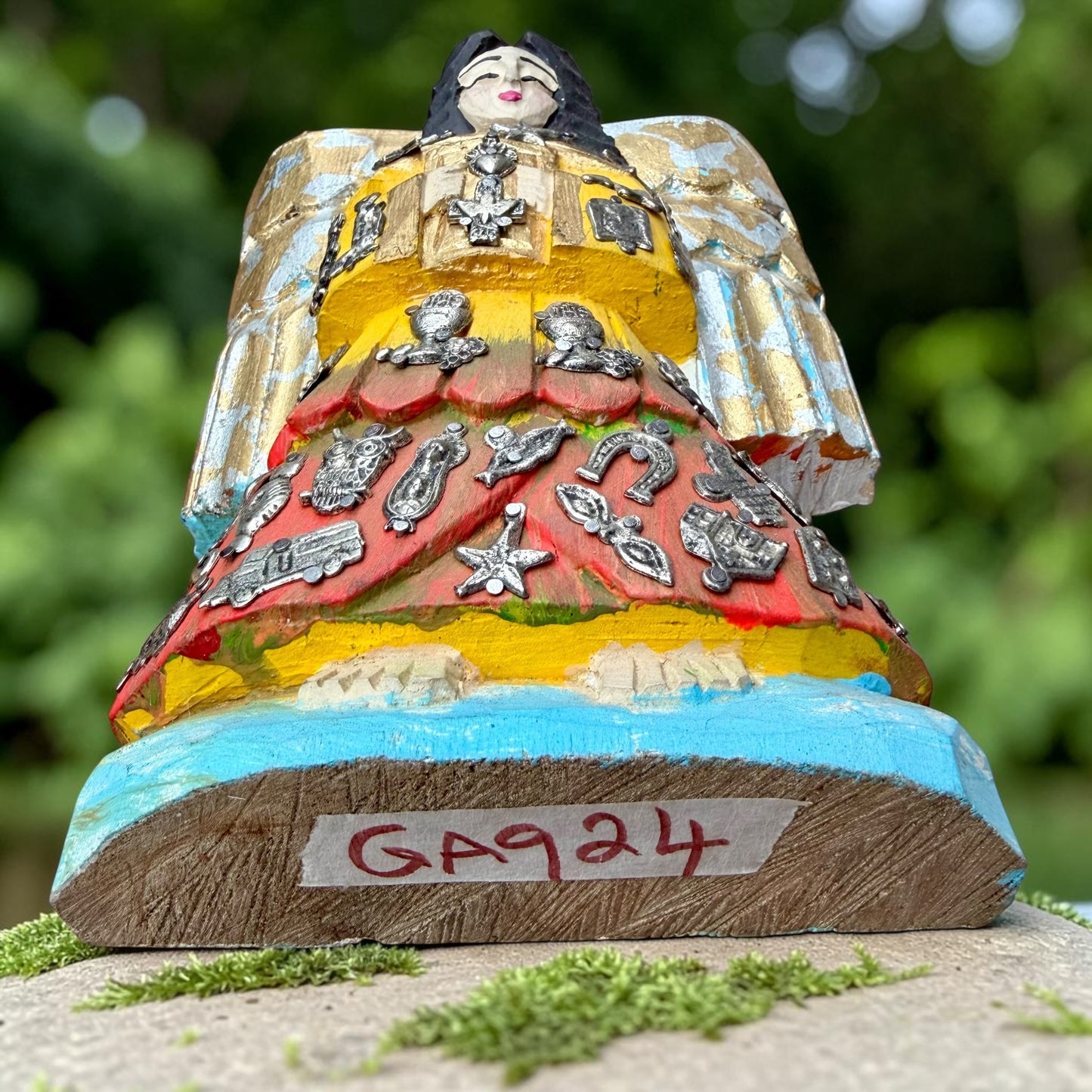 A handcrafted wooden Milagros angel figurine with large wings, decorated with metallic silver charms and painted in yellow, and orange, standing on a mossy stone with a blurred green garden background. Bottom view showing angel fingurine from slight incline.