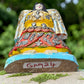 A handcrafted wooden Milagros angel figurine with large wings, decorated with metallic silver charms and painted in yellow, and orange, standing on a mossy stone with a blurred green garden background. Bottom view showing angel fingurine from slight incline.