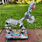 A hand-painted Mexican Talavera-style donkey figurine with a skeletal design, standing on a colorful floral base outdoors on a brick path with a green lawn in the background. Left side view of donkey