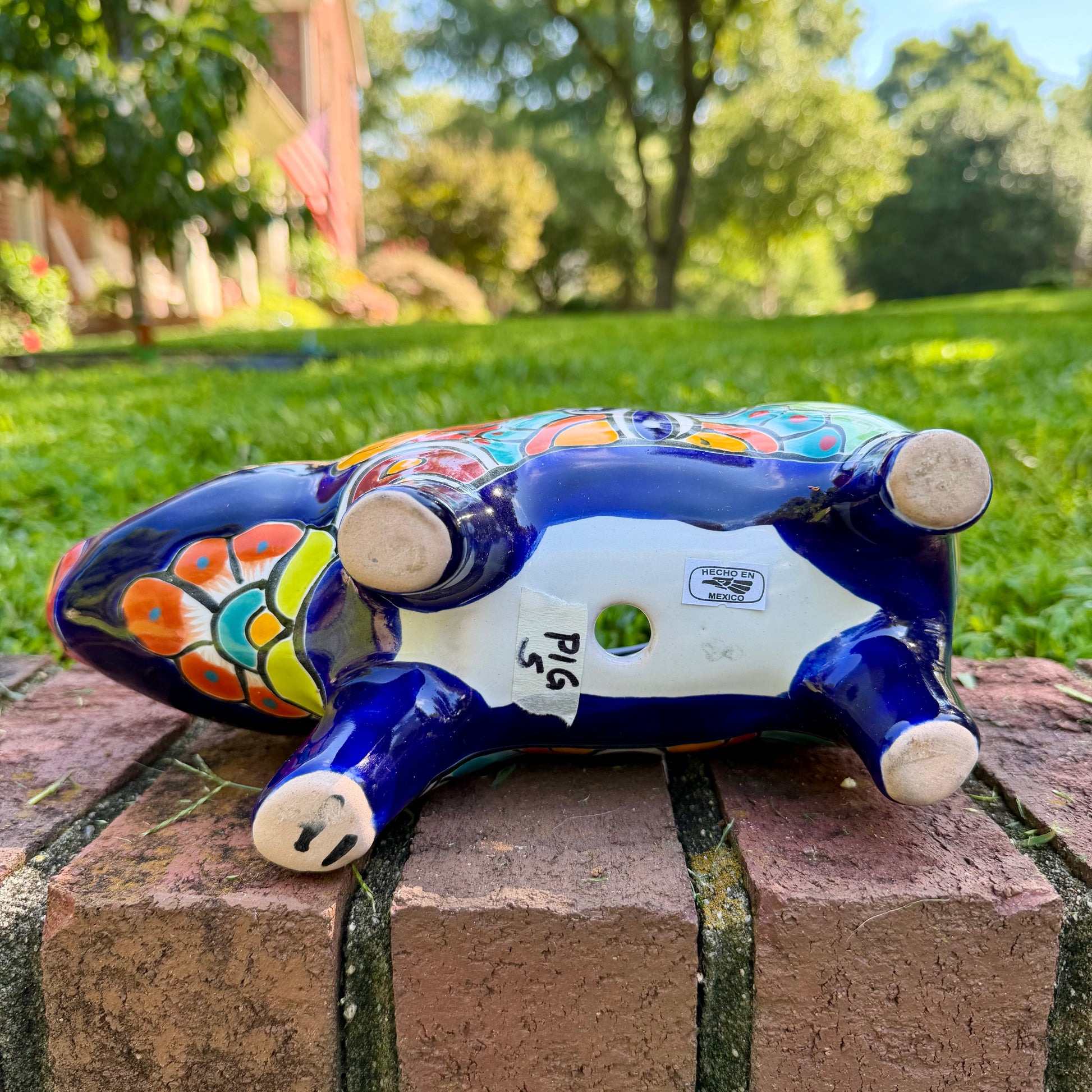 A glossy talavera pig figurine decorated with vibrant hand-painted patterns in blue, yellow, red, green, and white, standing on a brick surface with a blurred outdoor garden background, bottom drain hole.