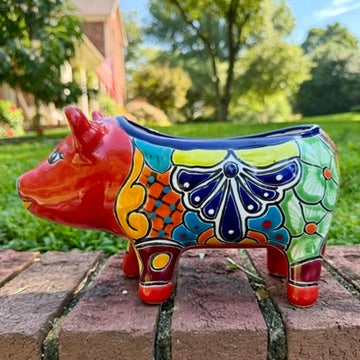 A colorful hand-painted red Talavera ceramic pig figurine standing on a brick surface with a blurred green lawn and trees in the background, right side view.
