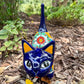 A colorful ceramic cat figurine with a glossy blue head and tail, yellow ears and paws, and a patterned body, standing outdoors on mulch with green plants in the background, Front face view.