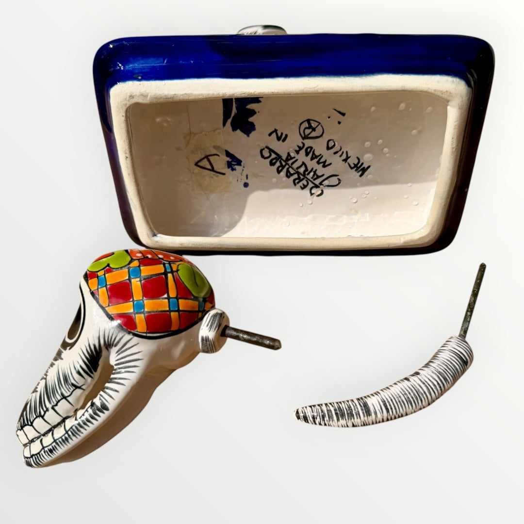 A talavera catrina dog skeleton figurine with colorful hand-painted floral and geometric patterns, standing on a matching decorated base against a plain white background. Bottom view of dog with head and tail separated.  Signature of artist on bottom.