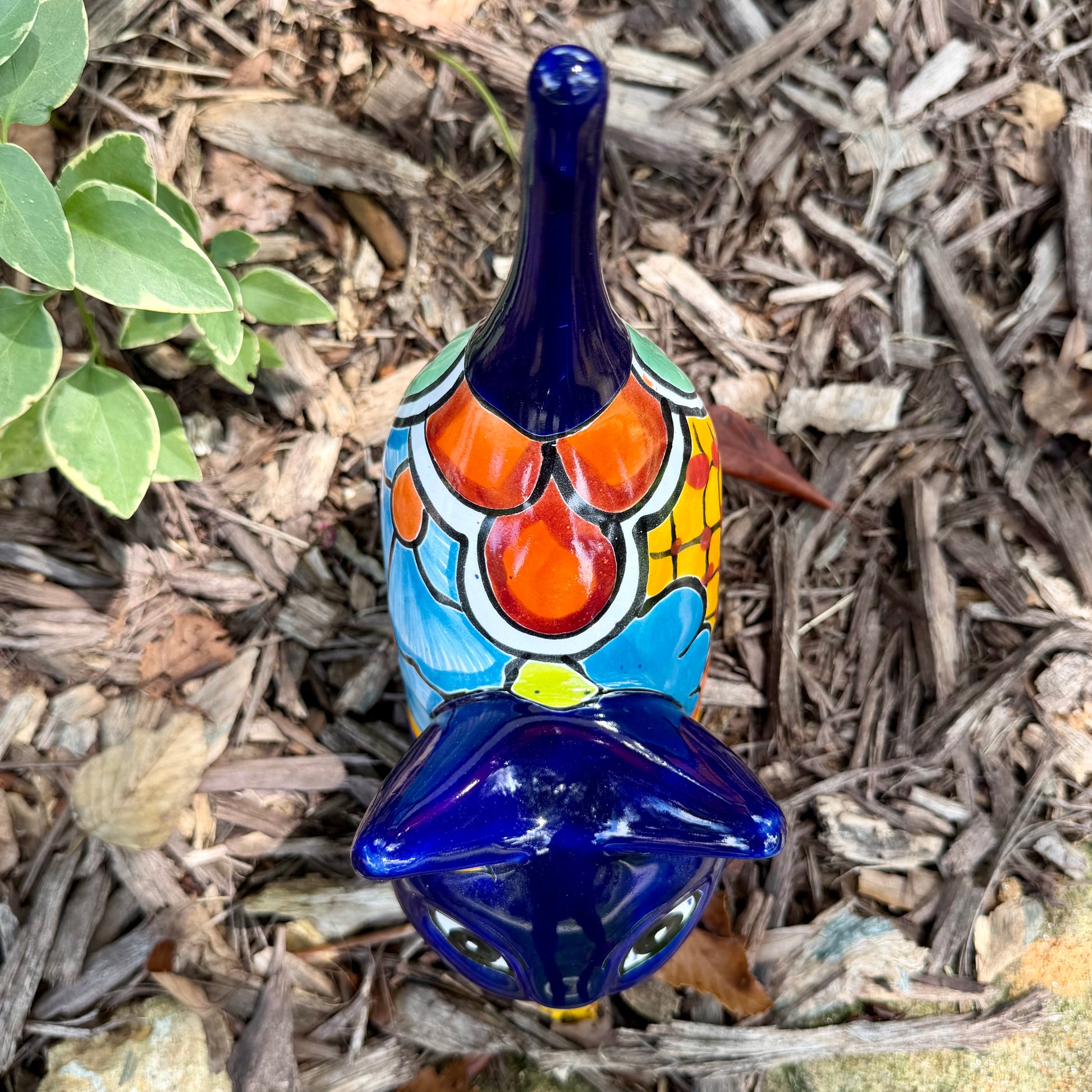 A colorful glazed ceramic cat figurine standing on mulch outdoors, with green plants blurred in the background. The cat is decorated with blue, orange, yellow, and white patterns, top view of cat.