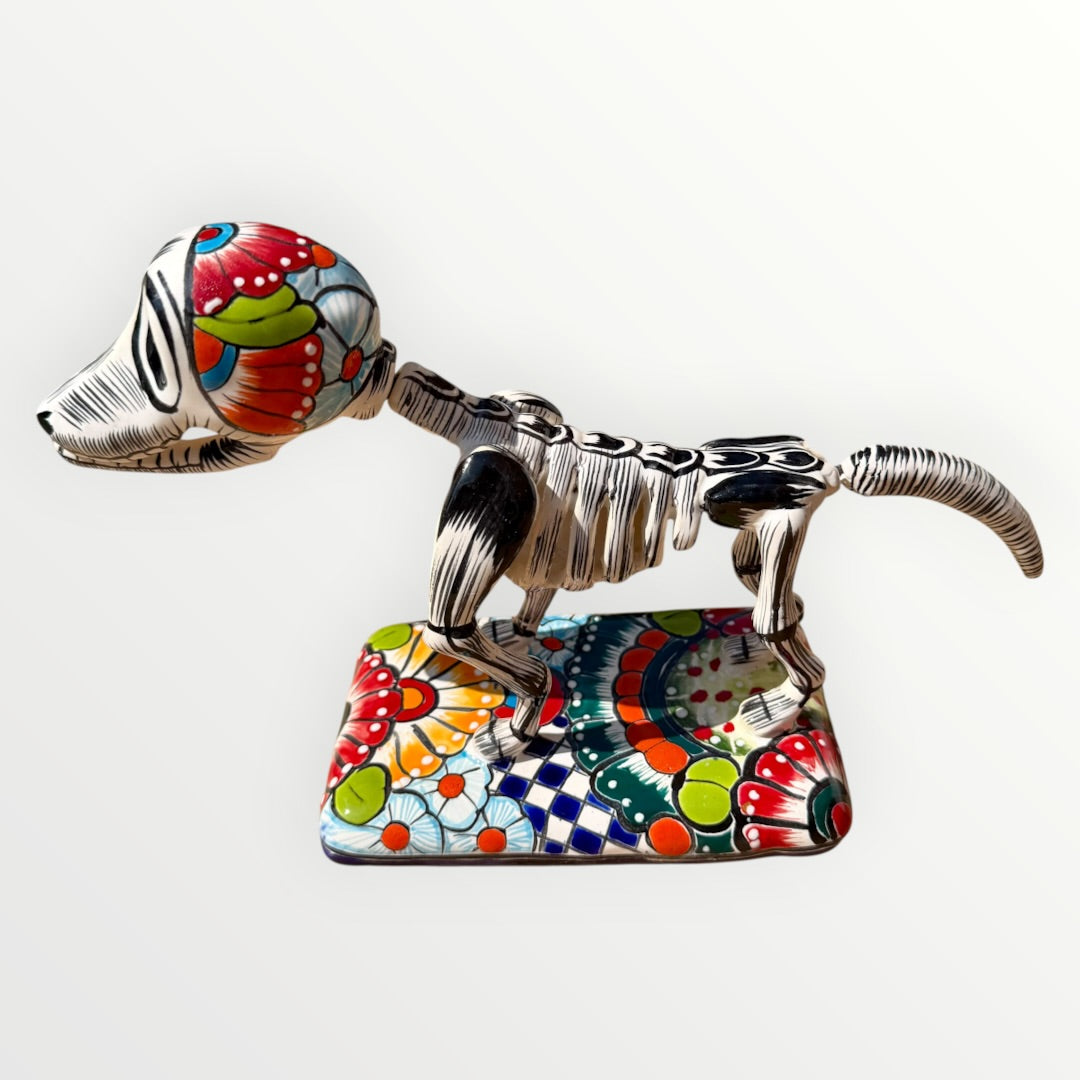 A ceramic Mexican-style skeleton dog figurine standing on a colorful floral base against a plain white background. Catrina right side view.